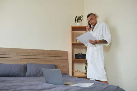 Serious young project manager in bathrobe making phone call to discuss report with colleague in the morningの写真素材
