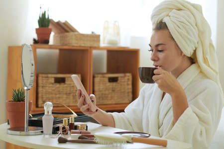 Young woman in bathrobe sitting at vanity, drinking coffee and reading news on smartphoneの写真素材