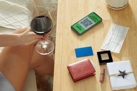 Smartphone with covid-19 vaccine passport, medical mask and wallet on table in front of woman drinking red wine a homeの写真素材
