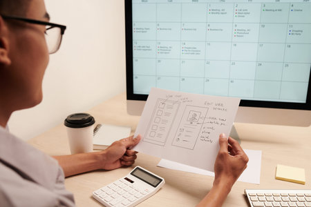 Project manager looking at hand drawn mockup of UI designer and filling schedule on computer