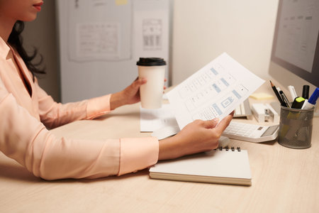 Pensive female UI designer drinking cup of coffee and looking at printed mockup of mobile...