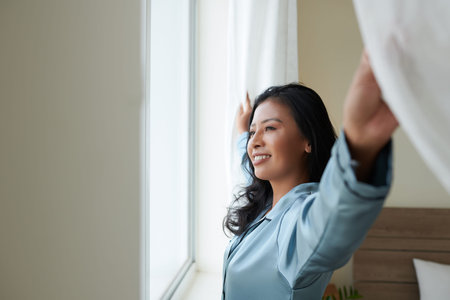 Happy young woman in silk loungewear opening curtains and looking through window enjoying new morningの写真素材