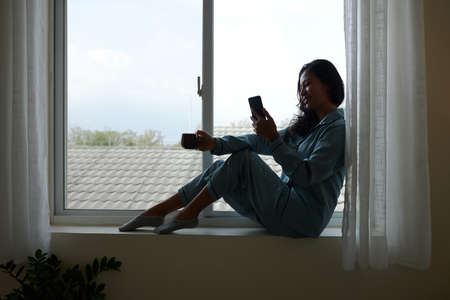 Happy young woman in pajamas sitting on window sill, drinking coffee or tea and reading news on smartphoneの写真素材