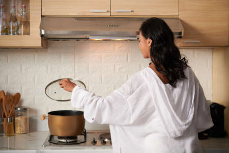 Young woman in loungewear opening lid of saucepan when cooking in kitchen, view from the backの写真素材