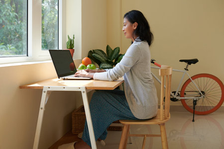 Smiling young woman with good posture wearing domestic clothes when working on laptop at homeの写真素材