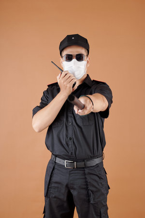 Security officer in protective mask talking on walkie-talkie and pointing at camera with baton