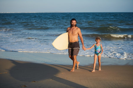 Smiling surfer walking on beach and holding hand of his little daughter