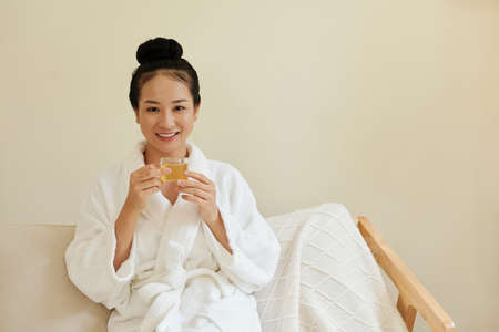 Portrait of smiling young Vietnamese woman in bathrobe holding cup of herbal tea and looking at cameraの写真素材