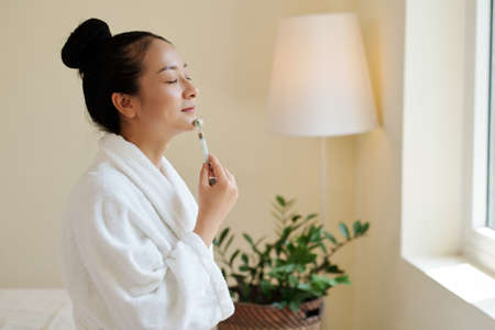 Smiling young woman in bathrobe massaging face with v-shape roller to stimulate blood flow and make skin look brighterの写真素材