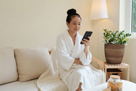 Positive young Vietnamese woman in bathrobe resting on couch after spa procedure and reading news on social mediaの写真素材