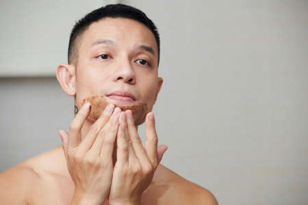 Asian young man looking at bathroom mirror and applying brown clay maskの写真素材