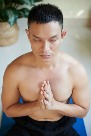Calm serious shirtless young man keeping hands in mudra gesture when meditating at home in the morningの写真素材