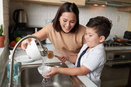Smiling mother helping son to rinse dishes under tap water after cleaningの写真素材