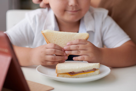 Cropped image of little boy eating tasty sandwich with ham and cheese