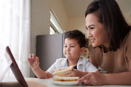 Proud boy eating sandwich and showing his school project on tablet computer to motherの写真素材