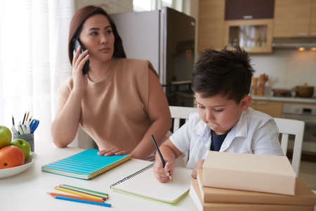Schoolboy doing homework under control of his mother talking on phone with teacherの写真素材