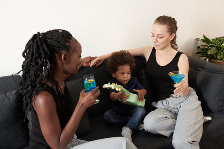 Diverse young couple drinking fruit cocktails when little son playing ukulele