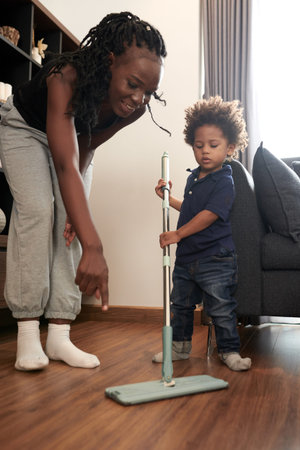 Young smiling mother teaching little son how to wipe wooden floor wit mop