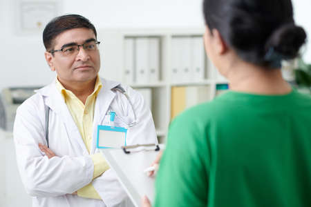 Pensive serious general practitioner with empty badge on labcoat talking to nurse filling medical formの写真素材