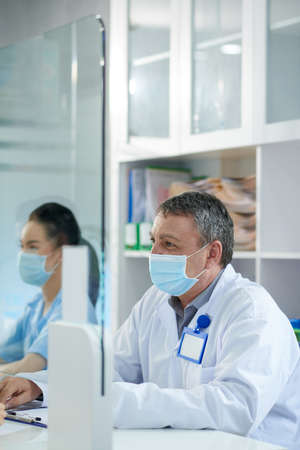 Senior doctor in medical mask working at clinic reception, consulting patients and filling formsの写真素材