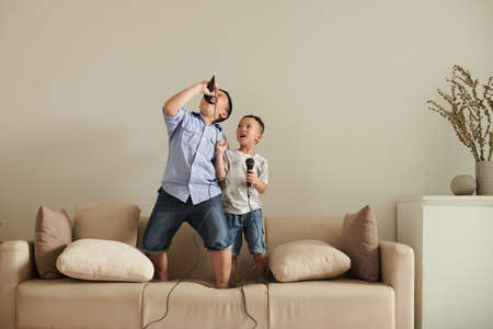 Two talented little brothers standing on sofa at home and singing into microphonesの写真素材