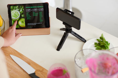 Plus-size female blogger checking green smoothie formula on tablet computer before filming cooking...