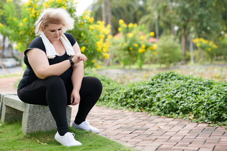 Plus-size young woman resting on bench and checking data on smartwatch after running in park in the...