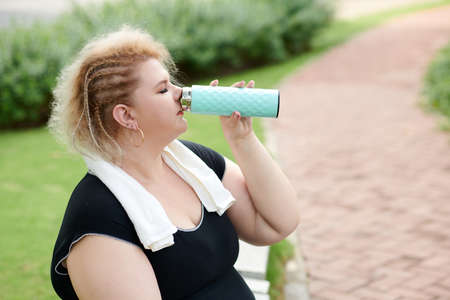Curvy stylish young woman drinking fresh water after jogging or working out in parkの写真素材