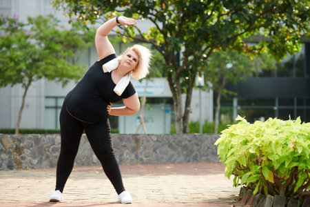 Plus-size young sportswoman doing side bends outdoor on sunny day