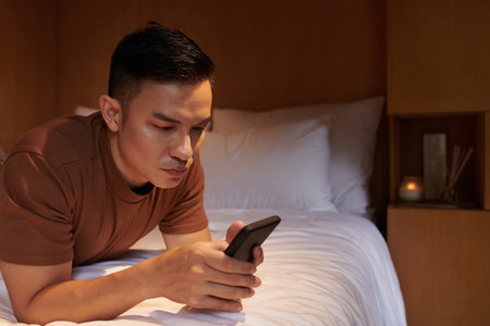 Serious young man resting on bed and answering text messages from his friends