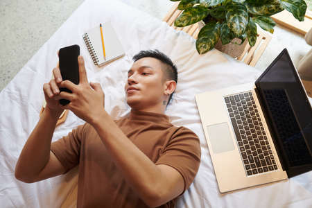 Yuong man lying on bed, texting friends or checking social media on smartphone instead of coding on laptopの写真素材