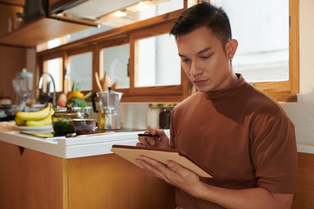 Serious young man standing in kitchen and checking grocery list on tablet computer