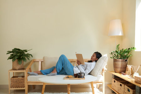 Young woman in comfy homewear relaxing on couch and reading good book