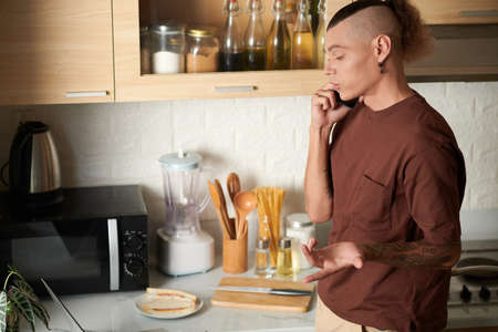 Young man talking on phone with colleague when cooking breakfast in kitchenの写真素材