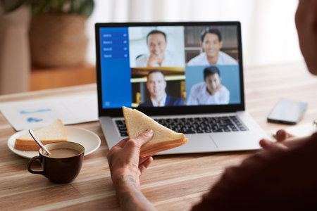 Young man eating sandwich with cup of coffee when having online meeting with colleagues