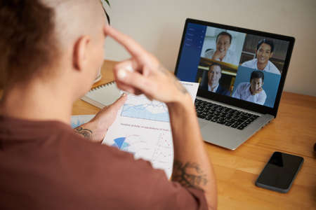 Pensive young entrepreneur analyzing chart with financial data and talking to colleague in online meetingの写真素材