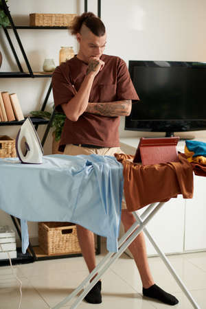 Pensive frowning young man watching webinar on tablet computer while ironing clothes at homeの写真素材