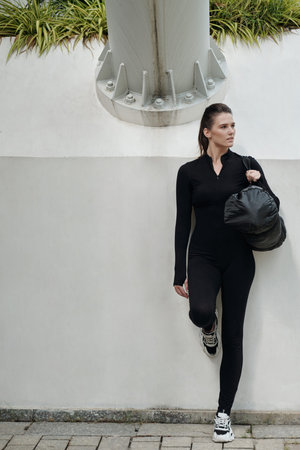 Young woman in dancewear holding gym bag and leaning on wall in park