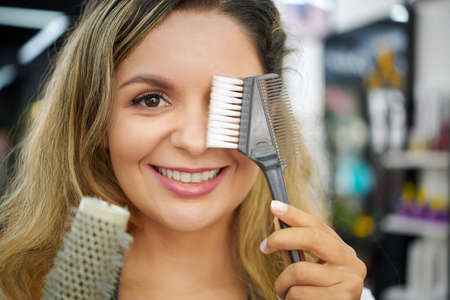 Face of smiling hair stylist covering one eye with plastic wide-toothed comb for hair dyeing and looking at cameraの写真素材