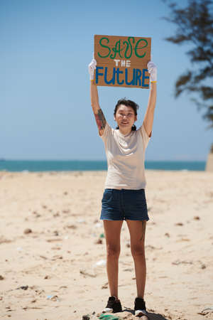 Happy excited young woman showing save the future placard when standing on dirty beachの写真素材