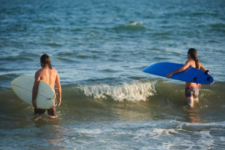 Surfers walking into ocean to catch waves in rays on setting sun