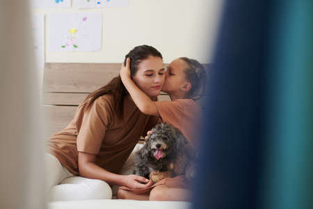 Girl kissing mother on cheek when they sitting on bed with small dogの写真素材