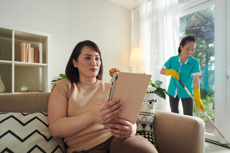 Woman enjoying reading e-book when professional cleaner wiping floor in backgroundの写真素材