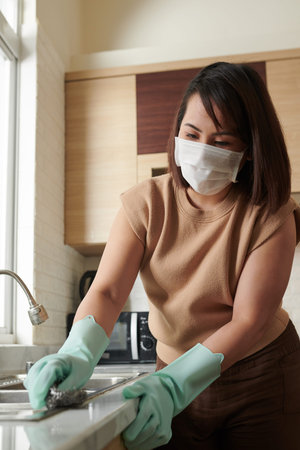 Woman wearing protective mask when cleaning kitchen sink with toxic detergent