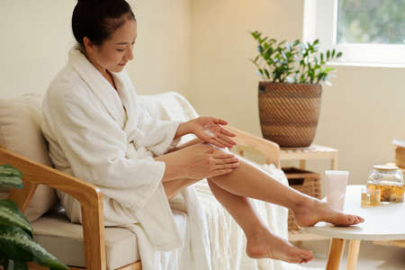 Young woman in soft bathrobe applying moisturizing lotion on legs after showerの写真素材