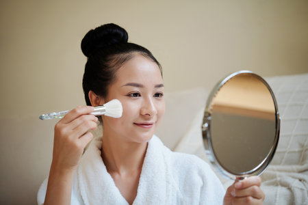 Portrait of happy young woman looking at table mirror when applying powder blushの写真素材