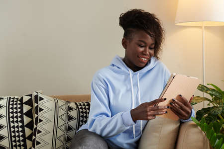 Portrait of positive young curly Black woman reading e-book when spending evening at homeの写真素材