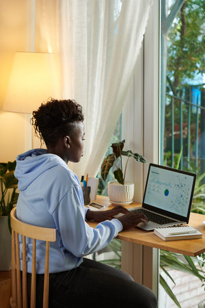 Concentrated young Black woman analyzing data in report on laptop when working from home
