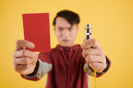 Frowning soccer referee showing red card and whistle, focus on foregroundの写真素材
