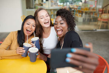 Joyful laughing young women taking photos on smartphone when sitting at cafe tableの写真素材